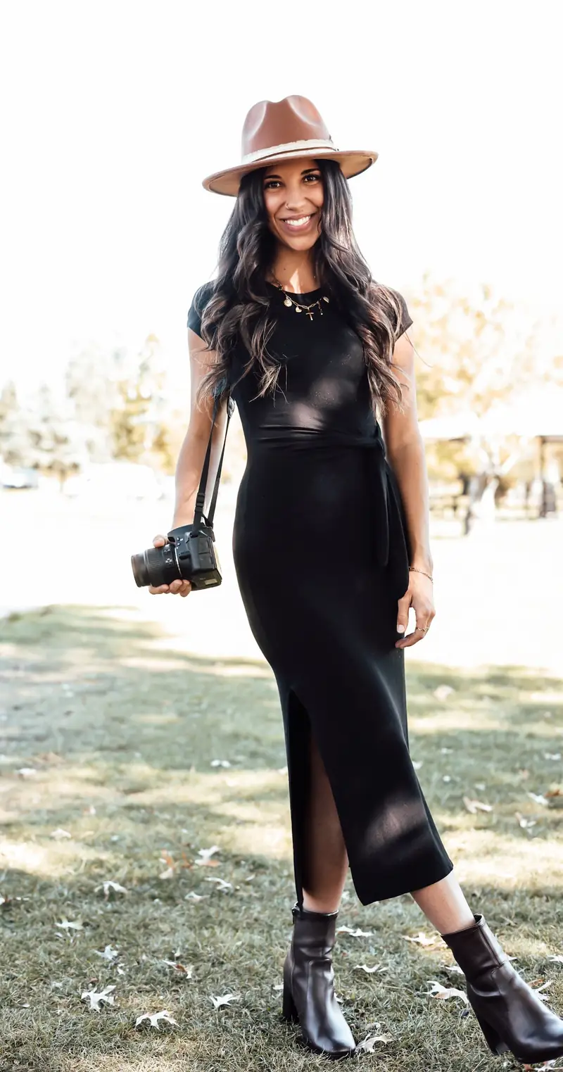 Alyssa standing in a park holding her camera, in a black dress, brown hat and ankle boots