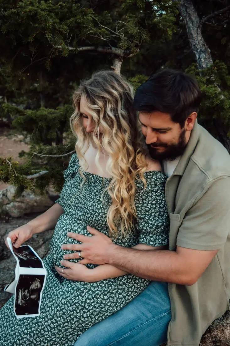 Expecting couple seated among pines holding an ultrasound strip