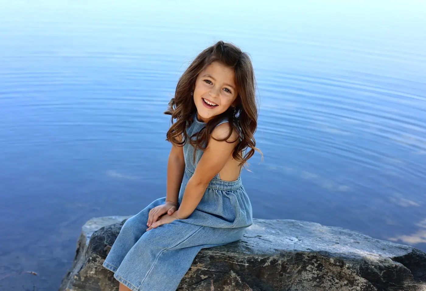 Smiling girl in a denim dress sitting on a rock beside calm blue water