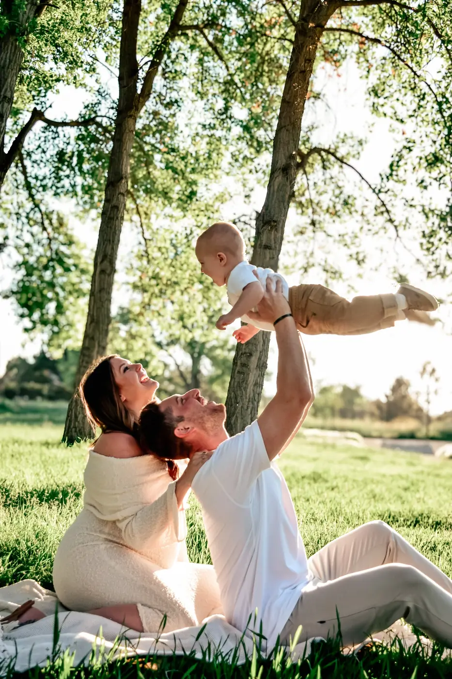 Dad lifting a laughing baby into the air while mom looks on, in a sunlit park