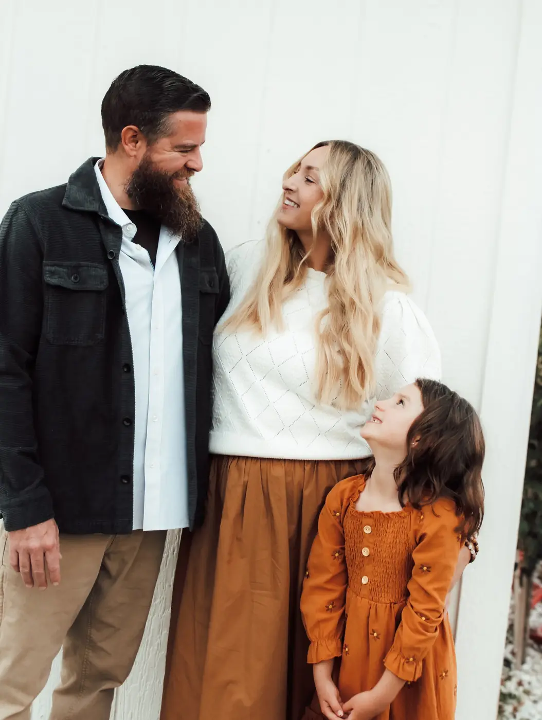 Mom, dad and daughter in rust and cream smiling at each other against a white wall