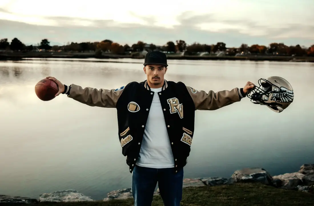 Senior in a varsity jacket holding a football and helmet at a lake